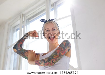 Sunny shot of joyful young pretty blonde woman with tattoos showing cherfully tongue to camera while demonstrating her strong biceps, posing in front of big window