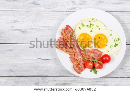 Sunny Side Up Eggs with crispy fried bacon, arugula and tomatoes on white plate on wooden table, view from above