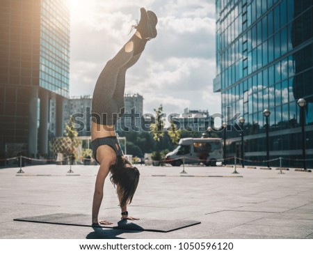 Sunny summer day. Young athletic woman doing handstand on city street among modern skyscrapers. Exercise outdoors, workout. Exercise for balance, yoga, training. Healthy lifestyle.