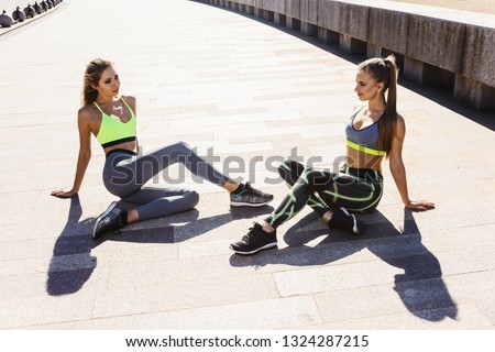 Sunny summer morning. Two young women athletes in sports clothes are sitting on the ground, relaxing after sports training workout and smiling. Happy healthy lifestyle concept
