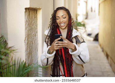 In a sunny urban alley, a cheerful young woman with long braided hair smiles at her smartphone, enjoying the warmth and reflecting the vibrant blend of technology and city life around her - Powered by Shutterstock