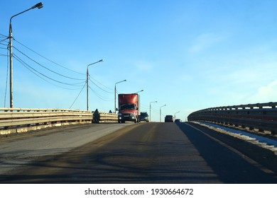 A Sunny Winter Day. Blue Sky. Cars Are Driving On A Transport Bridge. A Truck With A Red Cab Is Ahead. Lights, Wires, And The Bridge Fence Cast Shadows On The Road.