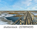 Sunny Winter day landscape of a long wooden boardwalk crossing a frozen march toward an ocean horizon at Grays Beach on Cape Cod, near Yarmouth, Massachusetts.