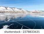 Sunny winter day on frozen Baikal Lake. Small Sea is covered with beautiful clear smooth blue ice with cracks. In distance snow-covered mountains and Sarma village. Beautiful winter landscape