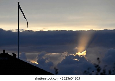 A sunray breaks through dark clouds rising towards the upper brighter sky. In the lower left, a part of a rooftop is seen with a flag pole in the background hoisting a flag.
