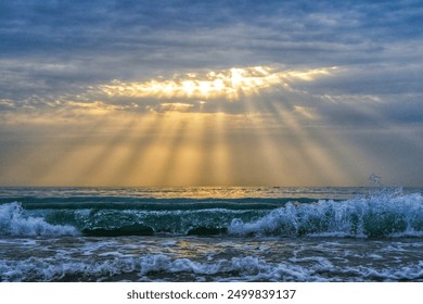 Sunrise between clouds, some sun rays can be seen coming out between them. Stormy morning, with waves in the foreground. - Powered by Shutterstock