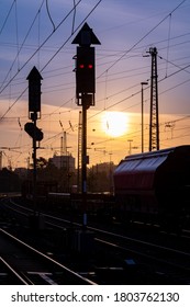 Sunrise With Glowing Rails At A Railway Station Near Hagen In North Rhine Westphalia Germany With A Passing Freight Train, Red Light Signals, Overhead Lines And Pylons