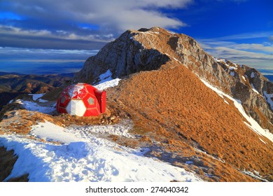 Sunrise illuminates mountain ridge and red refuge surrounded by snow patches - Powered by Shutterstock