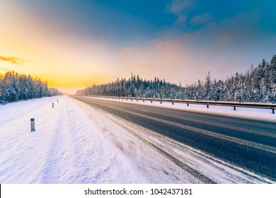 Sonnenaufgang an einem klaren Wintermorgen, eine leere Landstraße zum Schneefall durch einen Nadelwald. Aussicht von der Straßenseite. Russland, Europa. Schöne Natur. Orangenblauer Toneffekt.