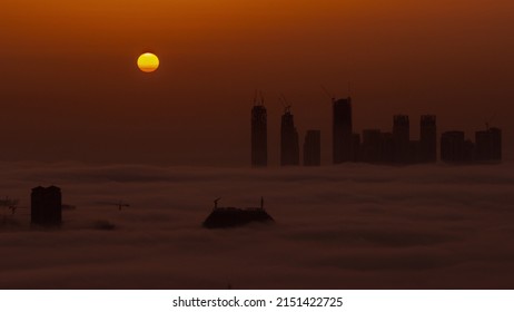 Sunrise Over Dubai Creek Harbor With Skyscrapers And Towers Covered By Morning Fog Aerial Timelapse. Big Red Sun Rise Up Behind Buildings And Orange Sky