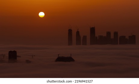 Sunrise Over Dubai Creek Harbor With Skyscrapers And Towers Covered By Morning Fog Aerial Timelapse. Big Red Sun Rise Up Behind Buildings And Orange Sky
