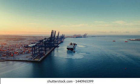 Sunrise over Felixstowe Container Port as two tugs shepherd a container ship from under blue gantry cranes with rows of brightly coloured shipping containers stacked behind them  - Shutterstock ID 1440965399
