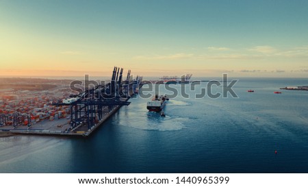 Sunrise over Felixstowe Container Port as two tugs shepherd a container ship from under blue gantry cranes with rows of brightly coloured shipping containers stacked behind them 