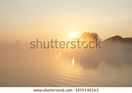 Sunrise over the foggy lake with the reflection of sun and trees in the water. Early fresh morning landscape. Mist on the water, forest silhouettes and the rays of the rising sun.