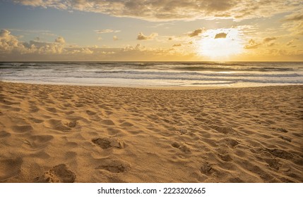 Sunrise Over Pacific Ocean At Cotton Tree, Maroochydore, Sunshine Coast. The Surf Is Rough And The Tide Coming In Onto A Sandy Beach With Lots Of Footprints. Cloudy Sky With A Golden Glow.