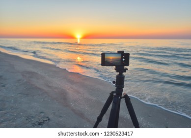 Sunrise over the sea coast. Fireball of the sun above the horizon in a colorful orange sky. Smartphone camera on a tripod in the foreground