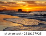 Sunrise over the water at Playa El Tunco, a popular surf beach in El Salvador. Golden light from the rising sun is seen behind the iconic rock formation and light is reflected off the sand and waves.