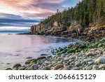 The sunrise paints the sky with color and shines warmly on the Otter Cliffs rising out of the sea at Boulder Beach in Acadia National Park, Mt. Desert Island, Maine.