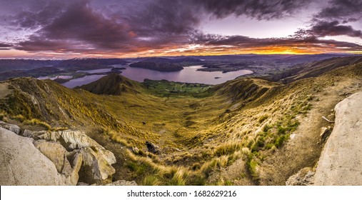 Sunrise panorama at Roys Peak summit, Wanaka, New Zealand. Colorful sky during dawn morning daybreak. At the top of Roys Peak track. View of cliffs, lake sounds and snowy Southern Alps mountain peaks.