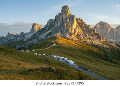 Sunrise at Passo Giau with road and parked cars and campers with sun shining at meadow and peaks in  the background during summer morning in august