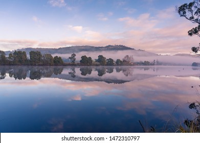 Sunrise Sky Reflected In Still Mirror Like River Water Surface With Misty Mountains On The Background. Huon River In Tasmania Nature Landscape. Eco Tourism In Australia