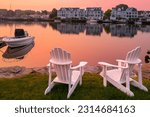 Sunrise, two white Adirondack chairs on the beach, a moored boat, and  wildfire smoke over the Mystic River marina village in Connecticut