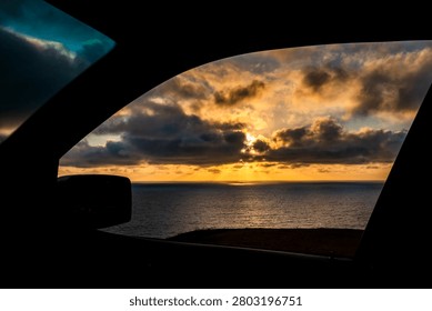 The sun's rays pierce through dramatic clouds, casting a golden glow on the vast ocean as seen from inside a vehicle