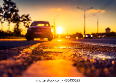 Sunset After Rain, The Car Parked On The Roadside And Headlights Of The Driving Cars On The Highway. Close Up View From The Level Of The Dividing Line, Image In The Orange-purple Toning