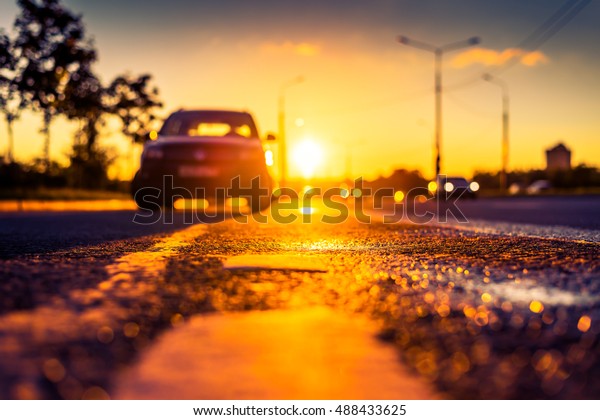 Sunset\
after rain, the car parked on the roadside and headlights of the\
driving cars on the highway. Close up view from the level of the\
dividing line, image in the orange-purple\
toning