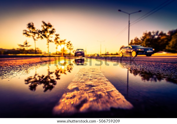 Sunset\
after rain, the car parked on the roadside and driving cars on the\
highway. Wide angle view of the dividing line level in a puddle,\
image vignetting and the orange-purple\
toning