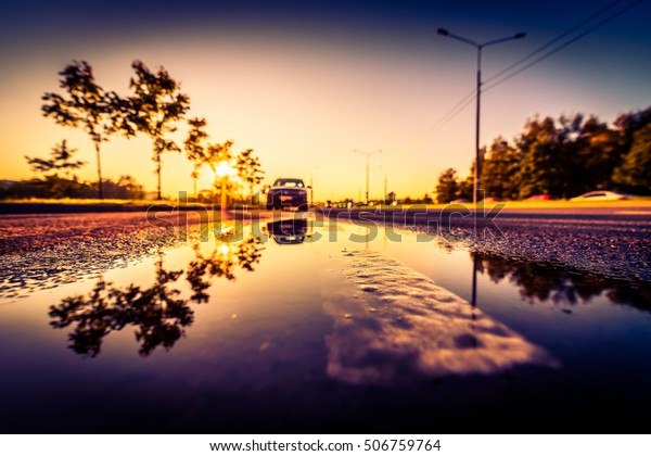 Sunset after rain, the car parked on the roadside\
and the headlights of the approaching cars. Wide angle view of the\
dividing line level in a puddle, image vignetting and the\
orange-purple toning
