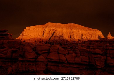 Sunset casts orange light on rock formation in desert. The landscape shows rough terrain and tall cliffs against a darkening sky.