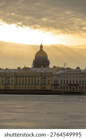 Sunset casts warm light on old buildings in Saint Petersburg during winter. The river reflects the colors of the sky. The dome of a notable structure stands tall against the evening light.