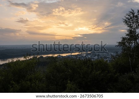 Sunset cloudy sky, bright sun over river, forest, industrial part of Ufa. Bashkiria. Greenery in foreground. Summer evening