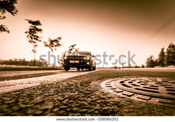 Sunset in the country, the car on the\
highway. Wide angle view of a close up from the level of the sewer\
manhole on pavement, image in the yellow\
toning