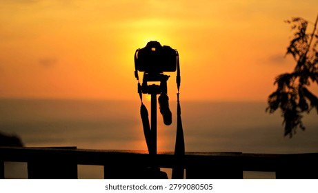 Sunset creates a beautiful silhouette of a camera on a tripod capturing the serene ocean view from a high vantage point