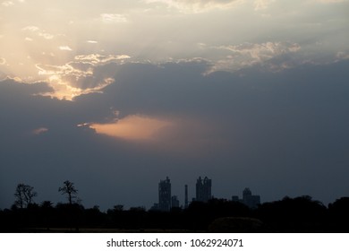 Sunset At Dramatic Sky With Dark Clouds Behind The Skyline Formed By The Cement Factory Of Chittorgarh, India - Sunrays Shine Through A Hole In The Clouds At Left Side Of The Skyline