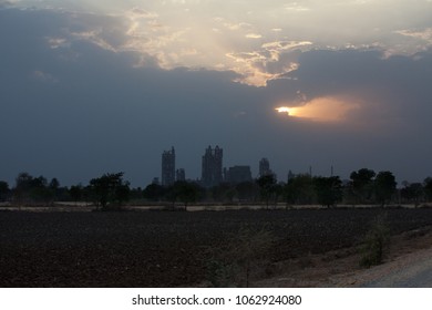 Sunset At Dramatic Sky With Dark Clouds Behind The Skyline Formed By The Cement Factory Of Chittorgarh, India - Sunrays Shine Through A Hole In The Clouds At Right Side Of The Skyline