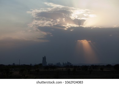 Sunset At Dramatic Sky With Dark Clouds Behind The Skyline Formed By The Cement Factory Of Chittorgarh, India - Sunrays Shine Through A Hole In The Clouds At Right Side Of The Building