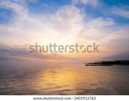 Sunset at the edge of a bay, fog at the landfall, pier and pilings in silhouette. Wispy clouds in blue sky, lightly rippling water.