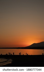 Sunset At The Foot Of The Beach In Marbella, Saying Goodbye To The Summer, With People Clustered Around The Shore Watching The Landscape.