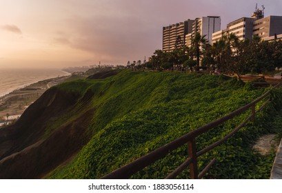 Sunset in front of cliff full of grass, trees, and big buildings.