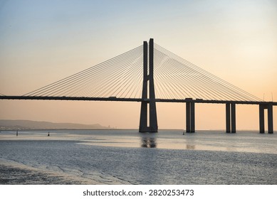 Sunset glows softly over vast bridge spanning calm waters. Silhouetted cables stretch from towering pylons to distant horizon. Gentle waves ripple beneath structure, reflecting golden light