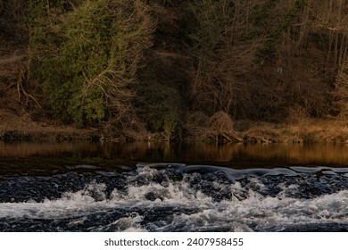 Sunset ignites river rapids, a symphony of motion. Rocks, clad in moss, bear witness to the dance of turbulent waters, illuminated by the warm palette of the departing day. - Powered by Shutterstock