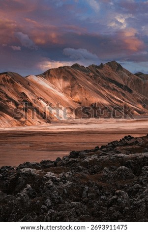 Sunset light casts warm hues on banded rhyolite mountains beyond a barren valley in Landmannalaugar, Iceland, with dark jagged lava rocks and moody clouds adding drama.