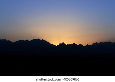 Sunset in mountains with gradient blue and orange sky and dark mountain peaks silhouette
