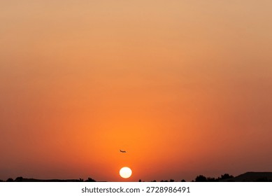 A sunset occurs in the sky while an airplane can be seen flying above the horizon. The sun glows as it lowers behind distant hills, creating a warm scene in the evening.