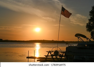 Sunset On A Lake With American Flag, Pontoon And Dock. 