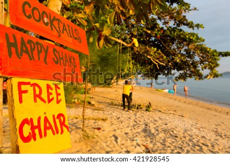 Sunset and orange saturated wooden signs standing on the beach and offering cocktails, happy shakes and free chairs with people enjoying in the background, Koh Chang Island. Thailand. Selective focus.