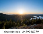 Sunset Over Acadia National Park A stunning sunset over the lakes and forests of Acadia National Park, Maine, with the sun setting behind a distant mountain range. The view from a mountaintop overloo